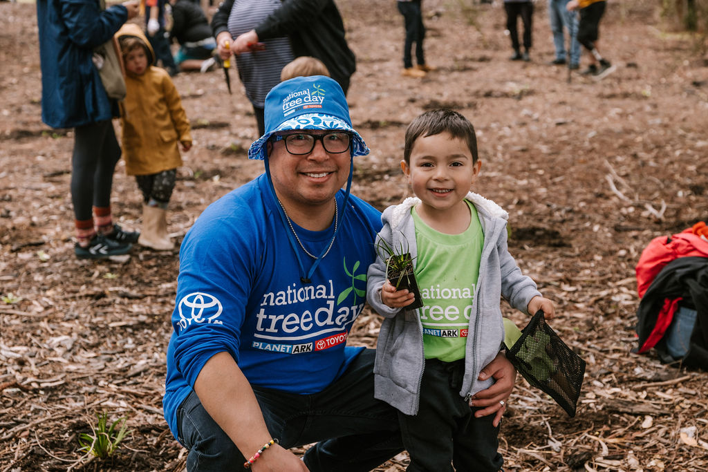 a man and a child wearing schools tree day merch, and the child is holding a plant ready to be planted in the ground